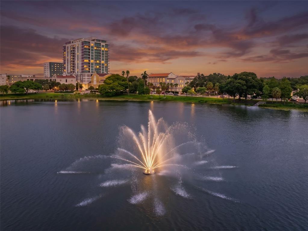 An illuminated water fountain sits in the center of a lake at sunset near buildings — 701 MIRROR LAKE DRIVE N UNIT 201, ST PETERSBURG, FL