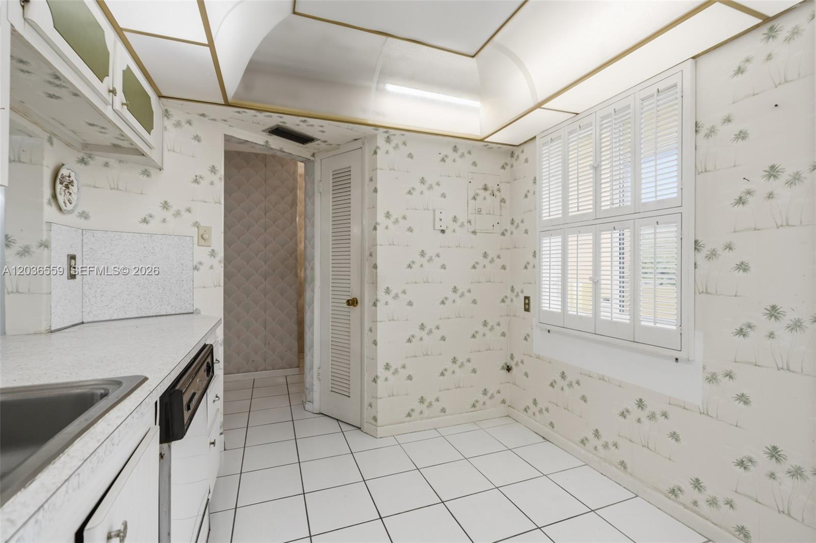 A kitchen features white tile flooring, patterned wallpaper, white cabinetry, and a louvered door — 1201 NE 191st St # G219, Miami FL 33179, MIAMI, FL