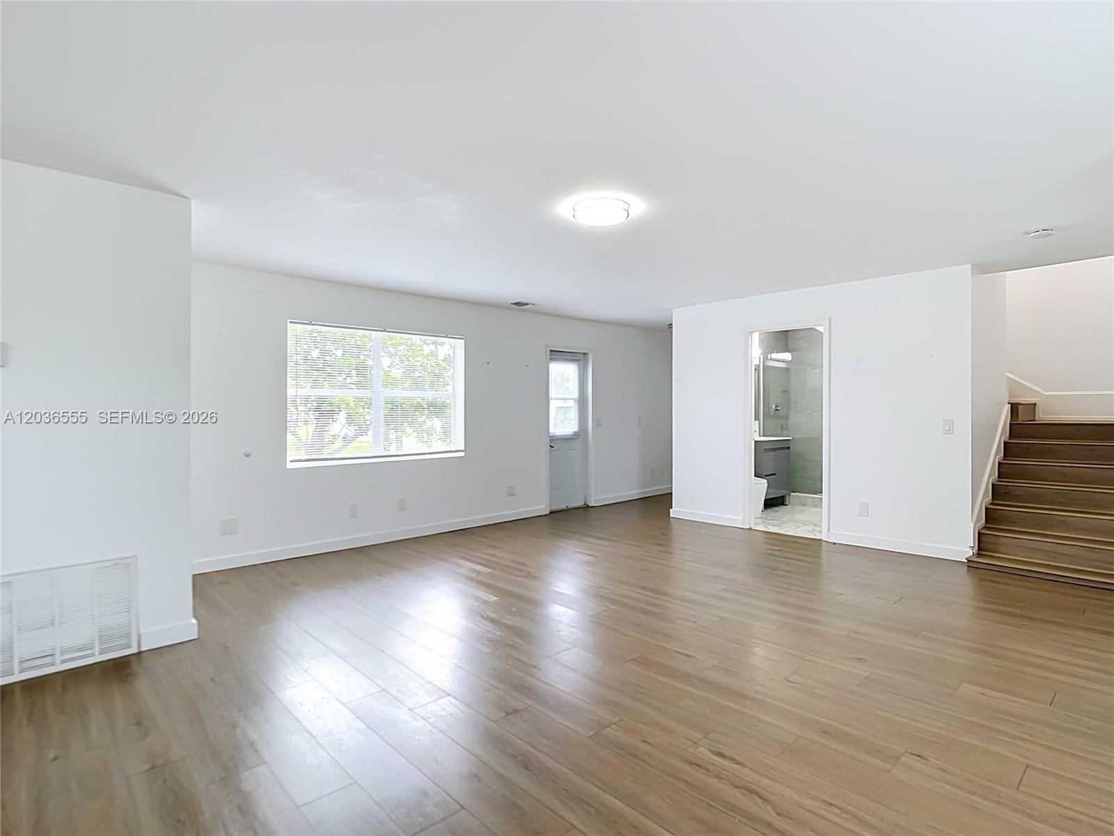 A bright, empty living room features light wood flooring, white walls, and a wooden staircase — 3303 6th, Lehigh Acres FL 33976, LEHIGH ACRES, FL
