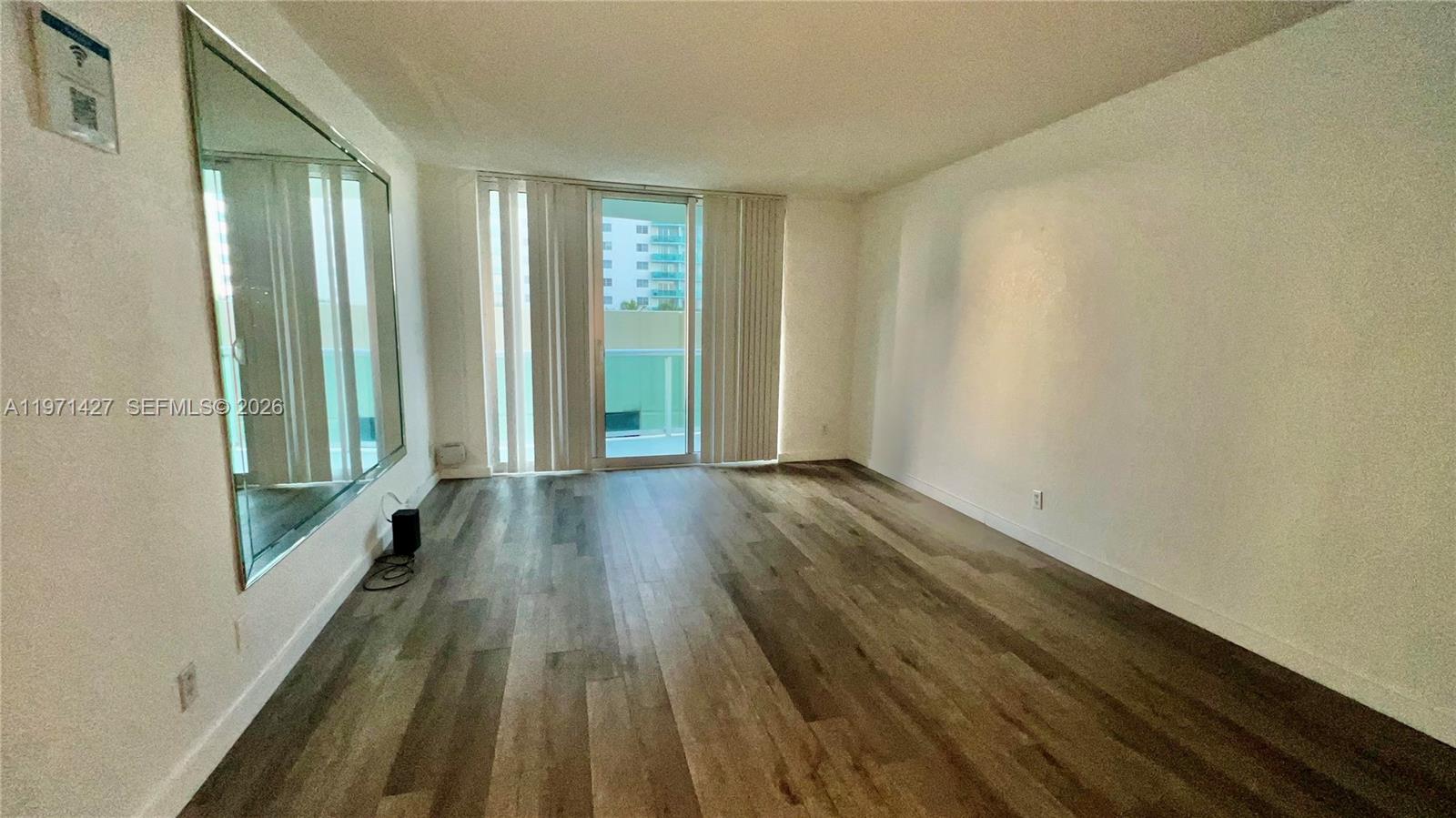 An empty living room features wood-look flooring, a large wall mirror, and a sliding glass door — 3901 S Ocean Dr # 3C, Hollywood FL 33019, HOLLYWOOD, FL