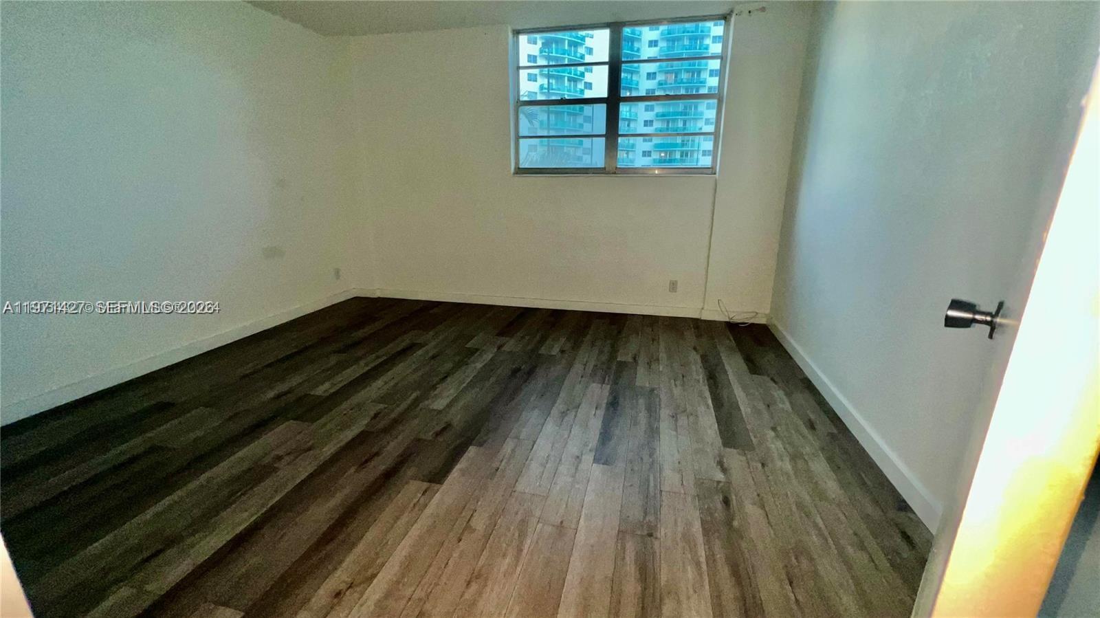 An empty room features white walls, dark wood-look flooring, and a window overlooking high-rise buildings — 3901 S Ocean Dr # 3C, Hollywood FL 33019, HOLLYWOOD, FL