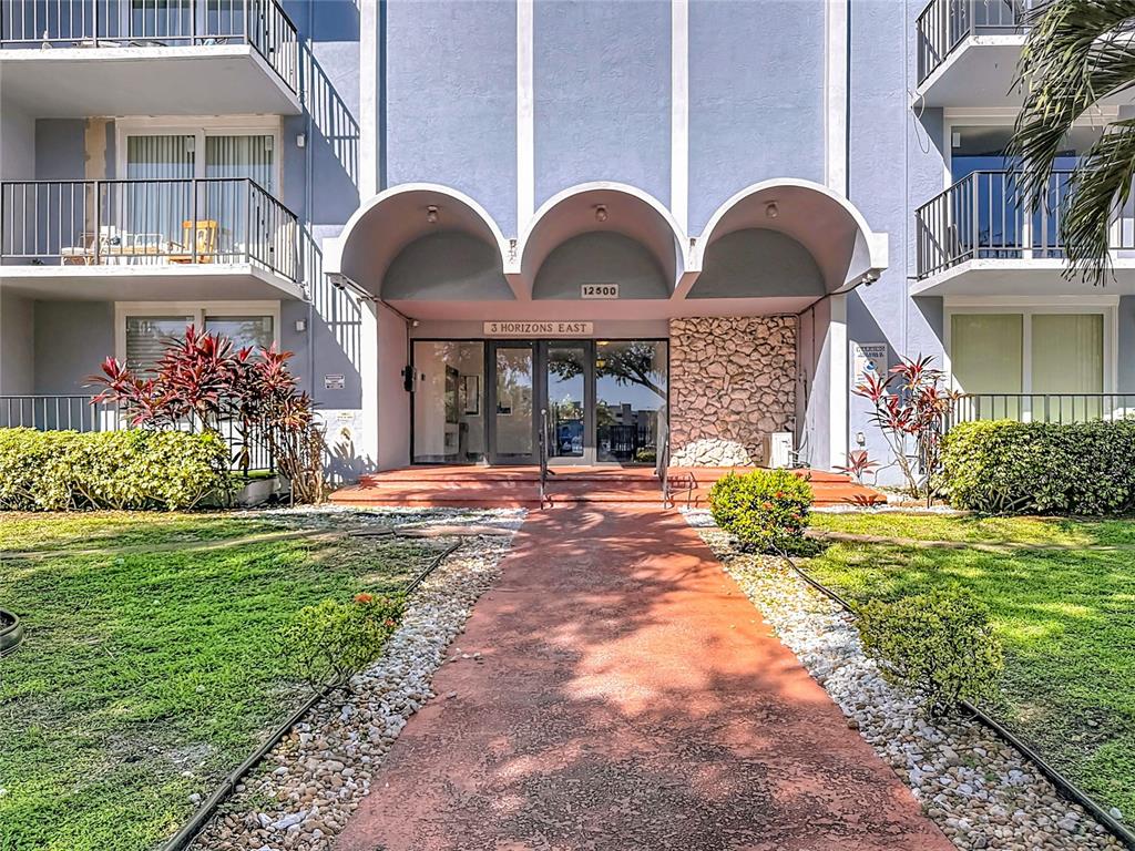 A red walkway leads to the entrance of a blue apartment building with arched overhangs — 12500 NE 15TH AVENUE UNIT 105, NORTH MIAMI, FL