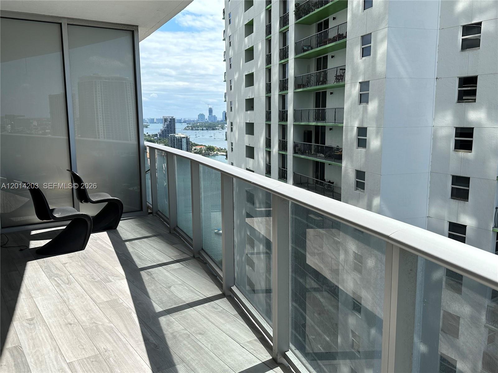 A balcony with wood-look tile flooring features two black chairs and a glass railing — 488 NE 18th St # 2502, Miami FL 33132, MIAMI, FL