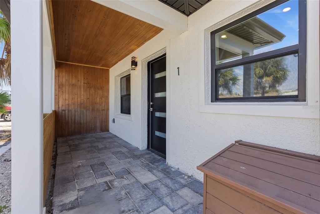 A covered porch with stone pavers, wood-paneled walls, and a modern black front door — 113 145TH AVENUE E UNIT 1, MADEIRA BEACH, FL