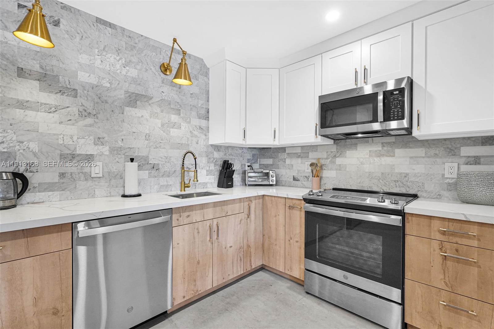 A kitchen features white upper cabinets, wood-toned lower cabinets, stainless steel appliances, and marble tile backsplash — 1012 NE 2nd Street, Hallandale Beach FL 33009, HALLANDALE BEACH, FL