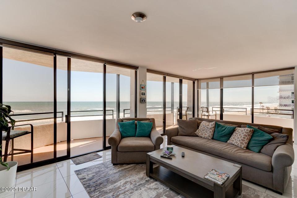 A living room with tile floors, a sofa, an armchair, and floor-to-ceiling windows overlooking the ocean — 2828 N ATLANTIC AVENUE UNIT 302, DAYTONA BEACH, FL