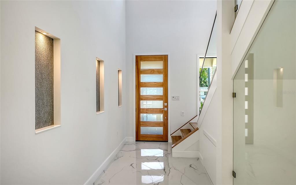 A modern entryway features white marble floors, a wooden front door, and recessed wall niches — 2483 BAY STREET, SARASOTA, FL