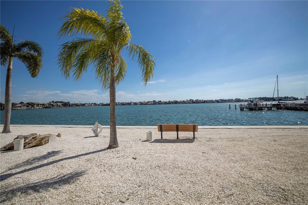 A bench sits on a shell-covered beach overlooking a bay with palm trees and boats — 5108 BRITTANY DRIVE S UNIT 204, ST PETERSBURG, FL