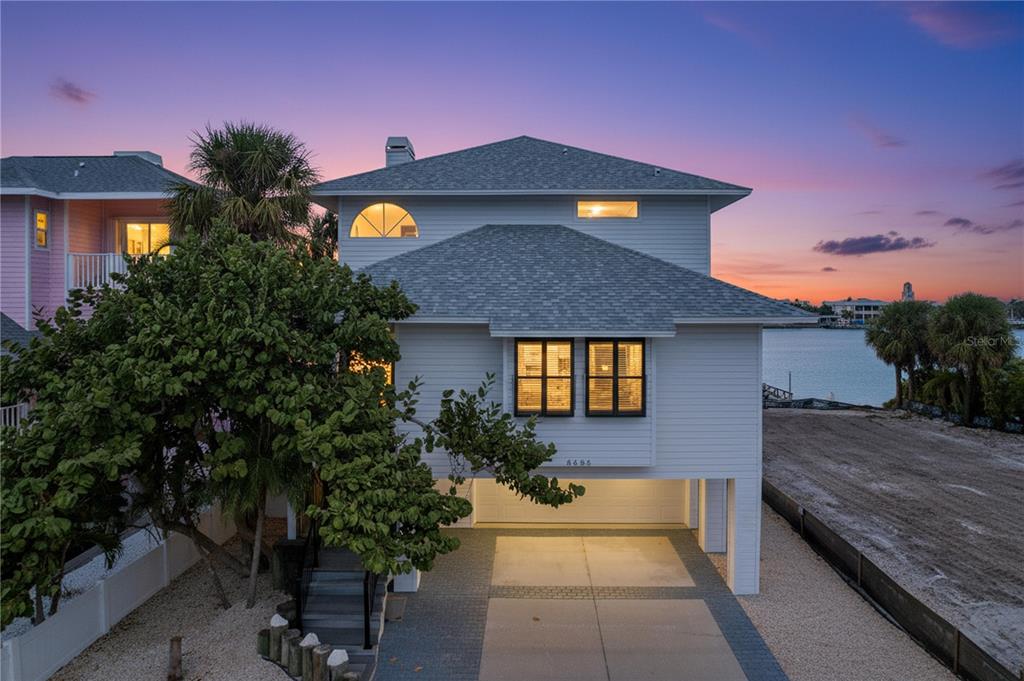 An elevated white house with a gray shingled roof stands beside a body of water — 8485 BLIND PASS DRIVE, TREASURE ISLAND, FL