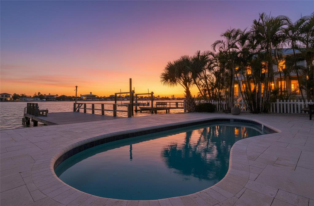 A swimming pool on a stone patio overlooks a wooden dock at sunset — 8485 BLIND PASS DRIVE, TREASURE ISLAND, FL