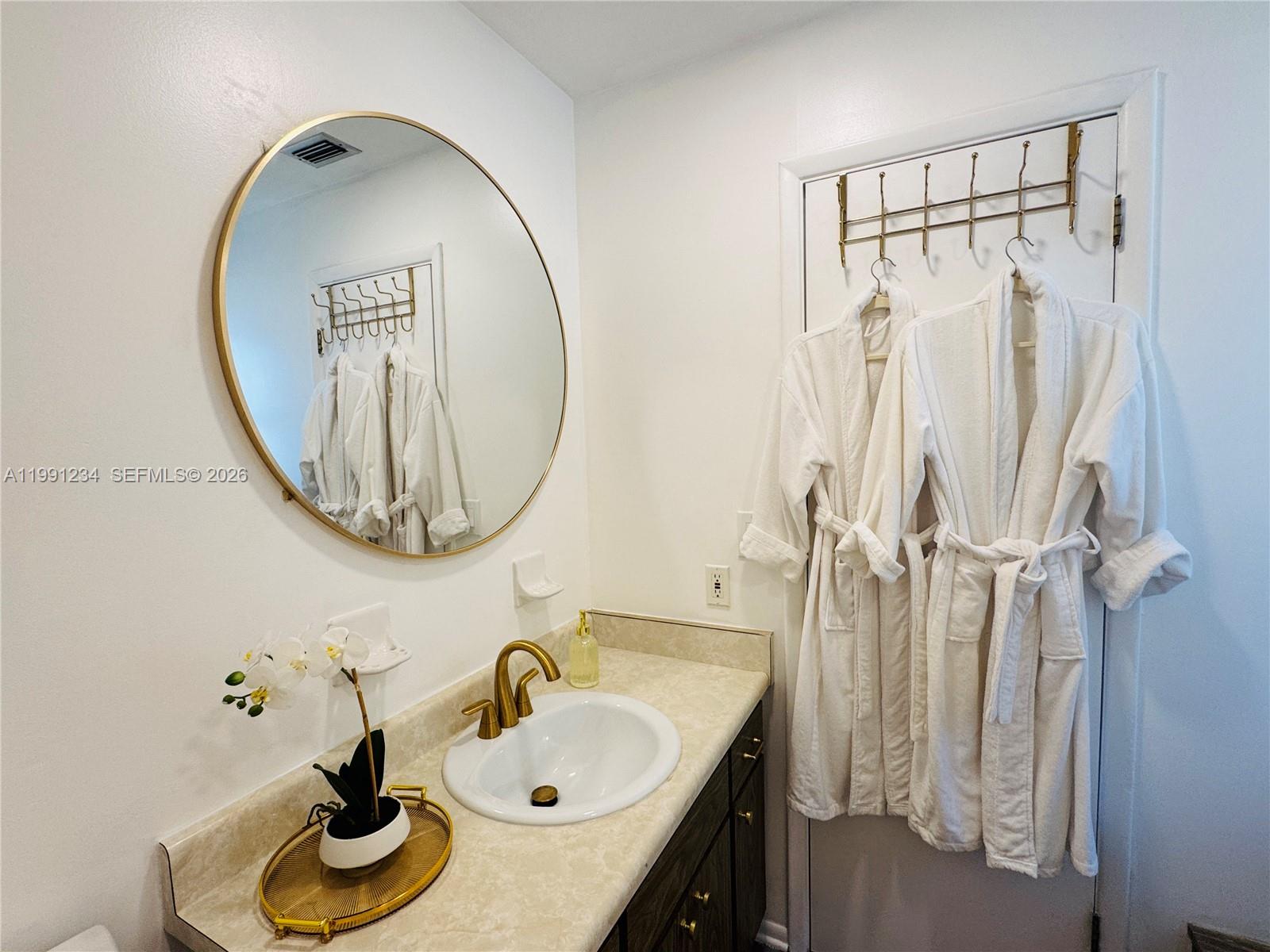 A bathroom features a vanity with a gold faucet, a round mirror, and two hanging robes — 5100 Rilma Ave # 0, Sarasota FL 34234, SARASOTA, FL