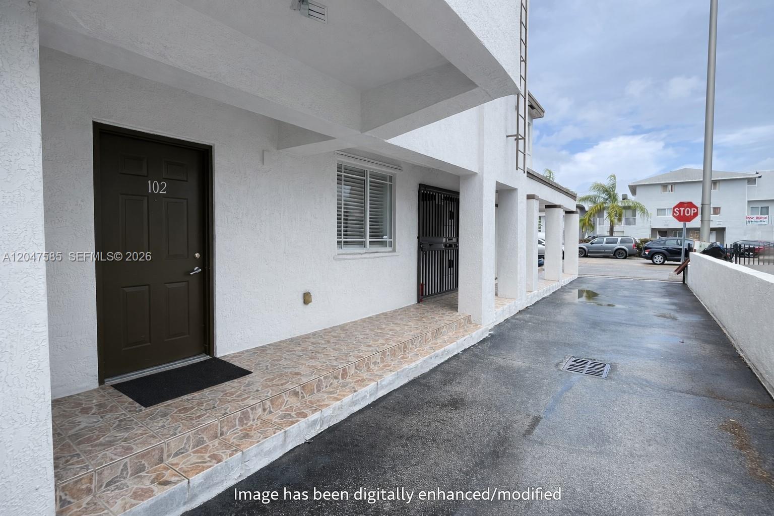 The exterior of a white building features a tiled porch, a brown door numbered 102, and a parking area — 318 E 6th St # 102, Hialeah FL 33010, HIALEAH, FL