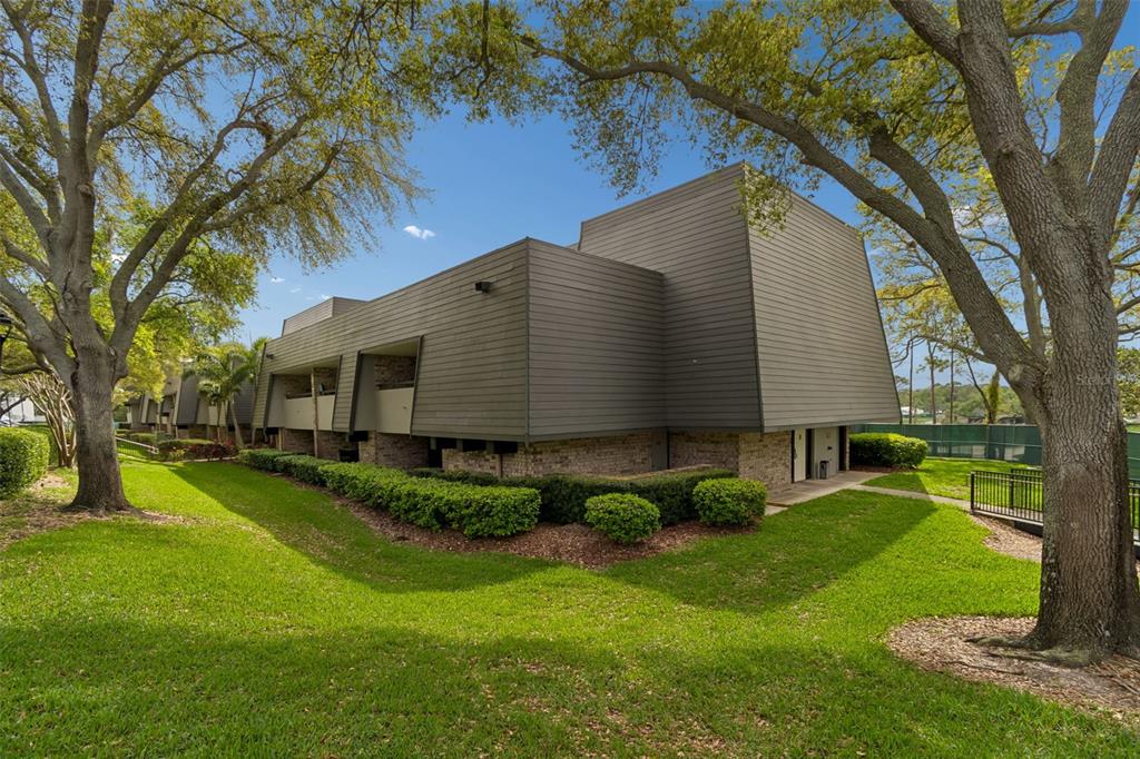 A multi-story building with dark horizontal siding and brick accents sits on a grassy lawn — 36750 US HIGHWAY 19 N UNIT 13118, PALM HARBOR, FL