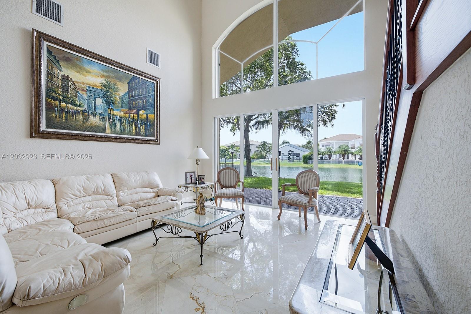 A living room features a large painting, white leather sofa, and floor-to-ceiling windows overlooking water — 18543 SW 49th St, Miramar FL 33029, MIRAMAR, FL