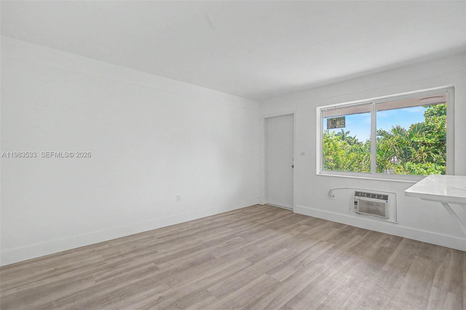 An empty room features white walls, light wood-look flooring, a window, and a wall-mounted air conditioner — 60 NW 79th St # 10, Miami FL 33150, MIAMI, FL