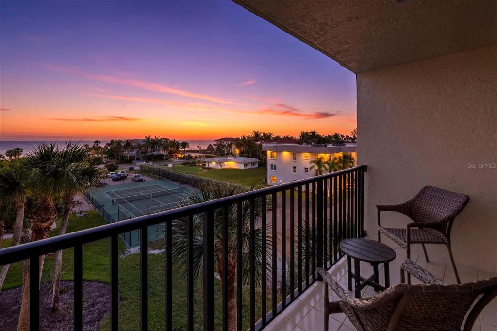 A balcony with wicker chairs overlooks a tennis court, palm trees, and a sunset ocean view — 3240 GULF OF MEXICO DRIVE UNIT B402, LONGBOAT KEY, FL