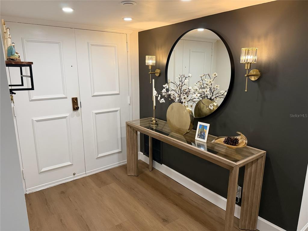 A foyer features a glass-topped console table, a round mirror, and wall sconces on a dark wall — 1480 GULF BOULEVARD UNIT 309, CLEARWATER BEACH, FL