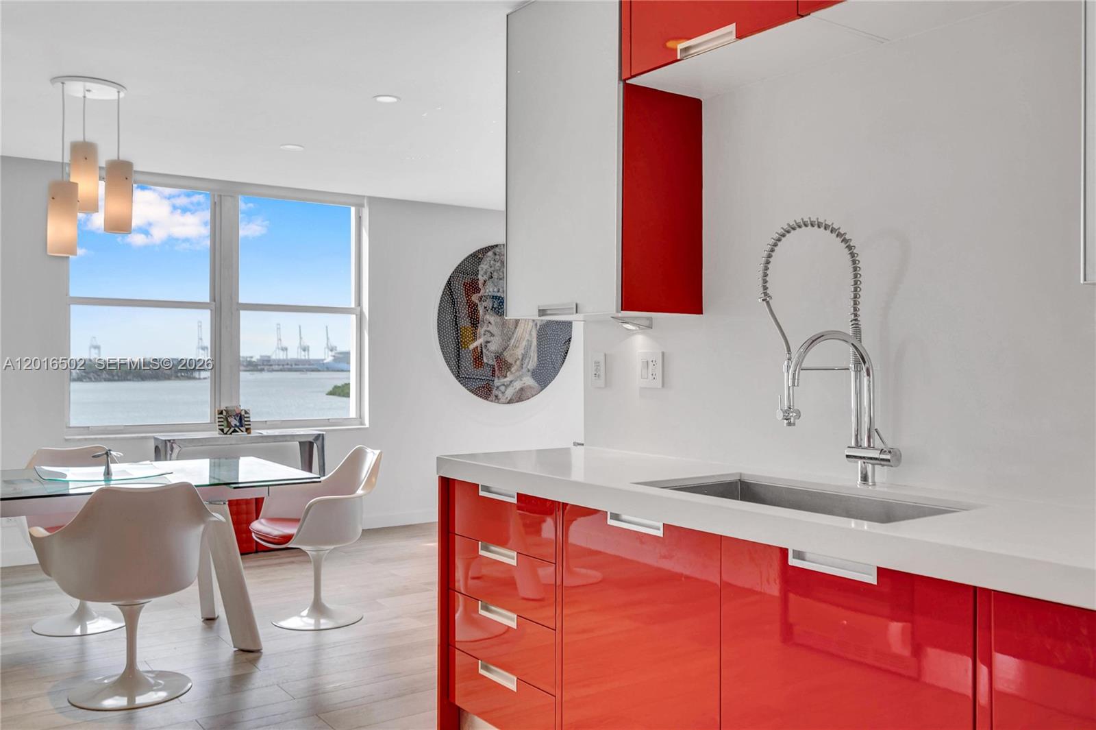 A modern kitchen with red cabinets and white countertops overlooks a dining area and water — 3 Island Ave # 08I, Miami Beach FL 33139, MIAMI BEACH, FL