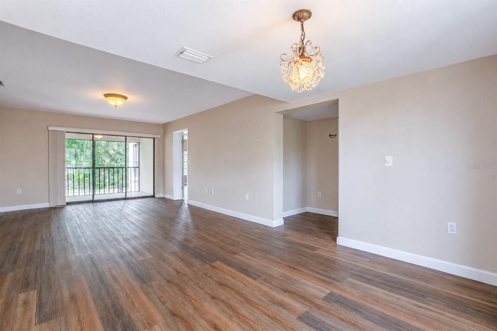 An empty living room features wood-look flooring, a crystal chandelier, and a sliding glass door — 2497 HERON TERRACE UNIT 203C, CLEARWATER, FL