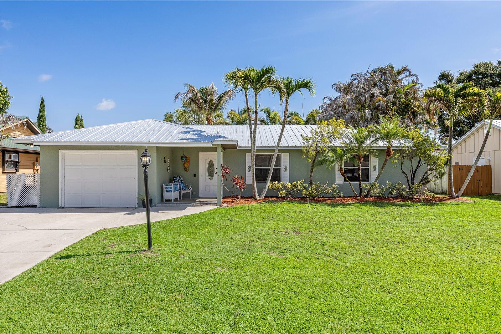 A single-story house with a white garage door, metal roof, and palm trees in front — 687 NE Maranta Terrado, Jensen Beach FL 34957, JENSEN BEACH, FL