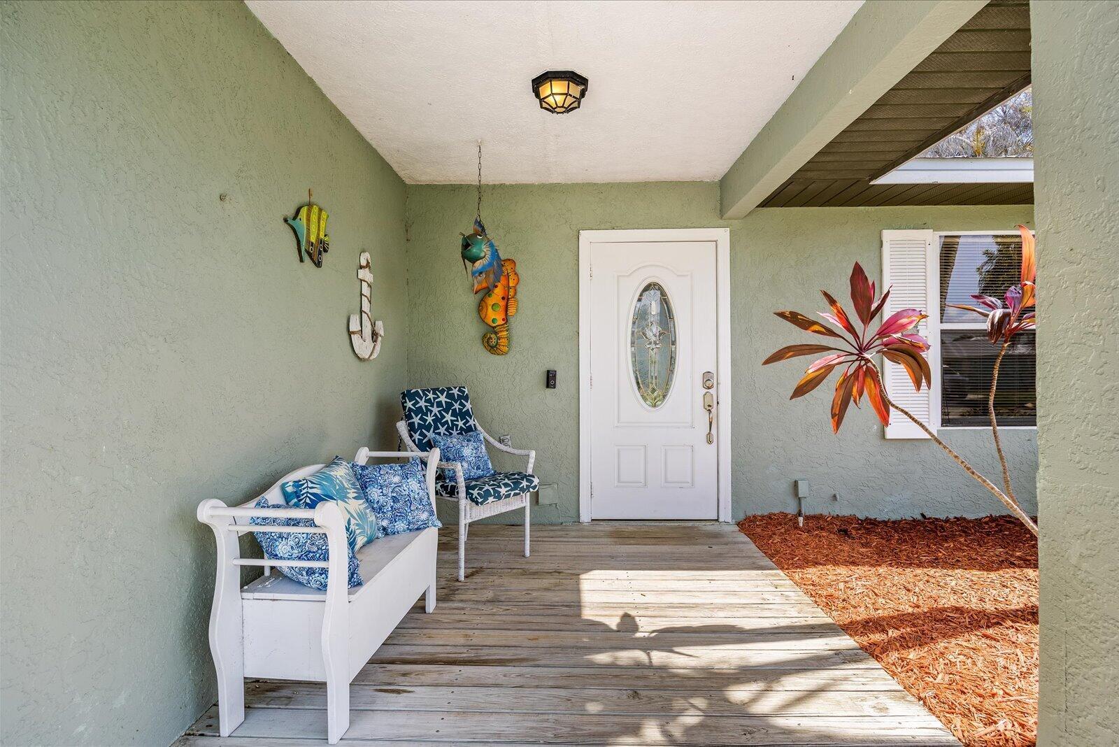 A covered porch features a wooden deck, white bench, wicker chair, and a front door — 687 NE Maranta Terrado, Jensen Beach FL 34957, JENSEN BEACH, FL