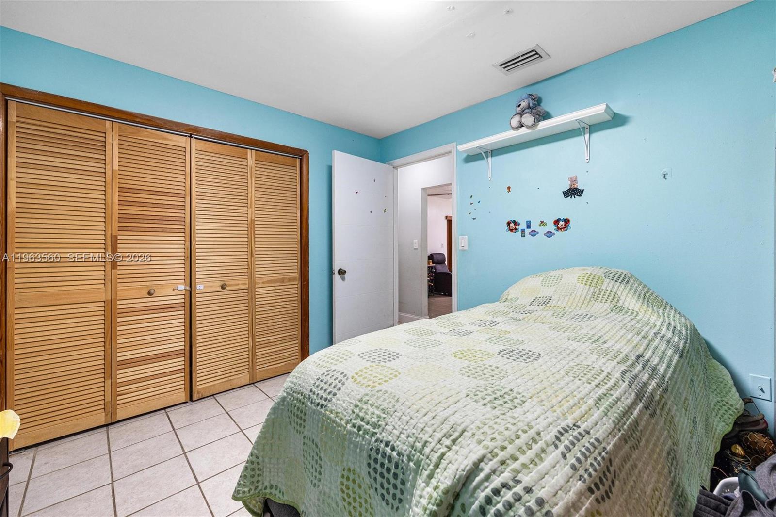 A bedroom features light blue walls, a bed with a patterned quilt, and louvered closet doors — 14217 SW 102nd St, Miami FL 33186, MIAMI, FL