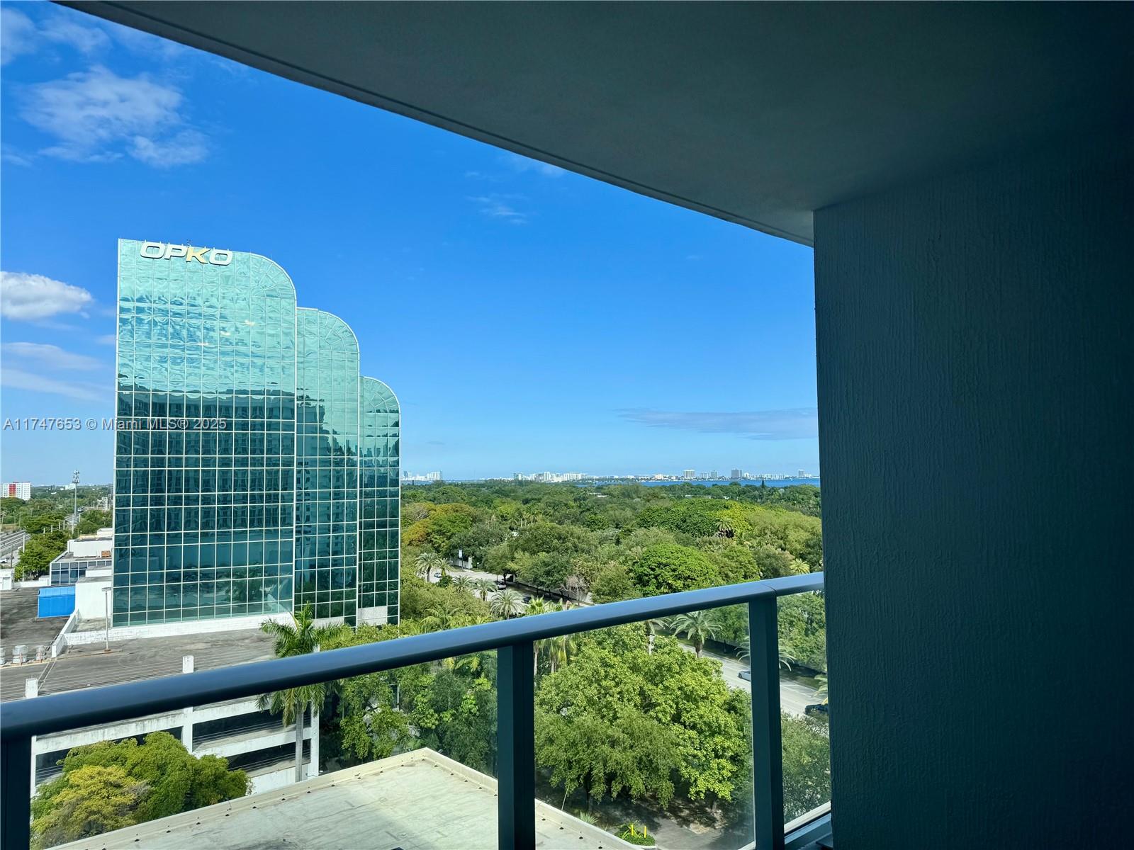 A balcony view overlooks a glass office building, lush green trees, and a distant bay — 4250 Biscayne Blvd # 1006, Miami FL 33137, MIAMI, FL