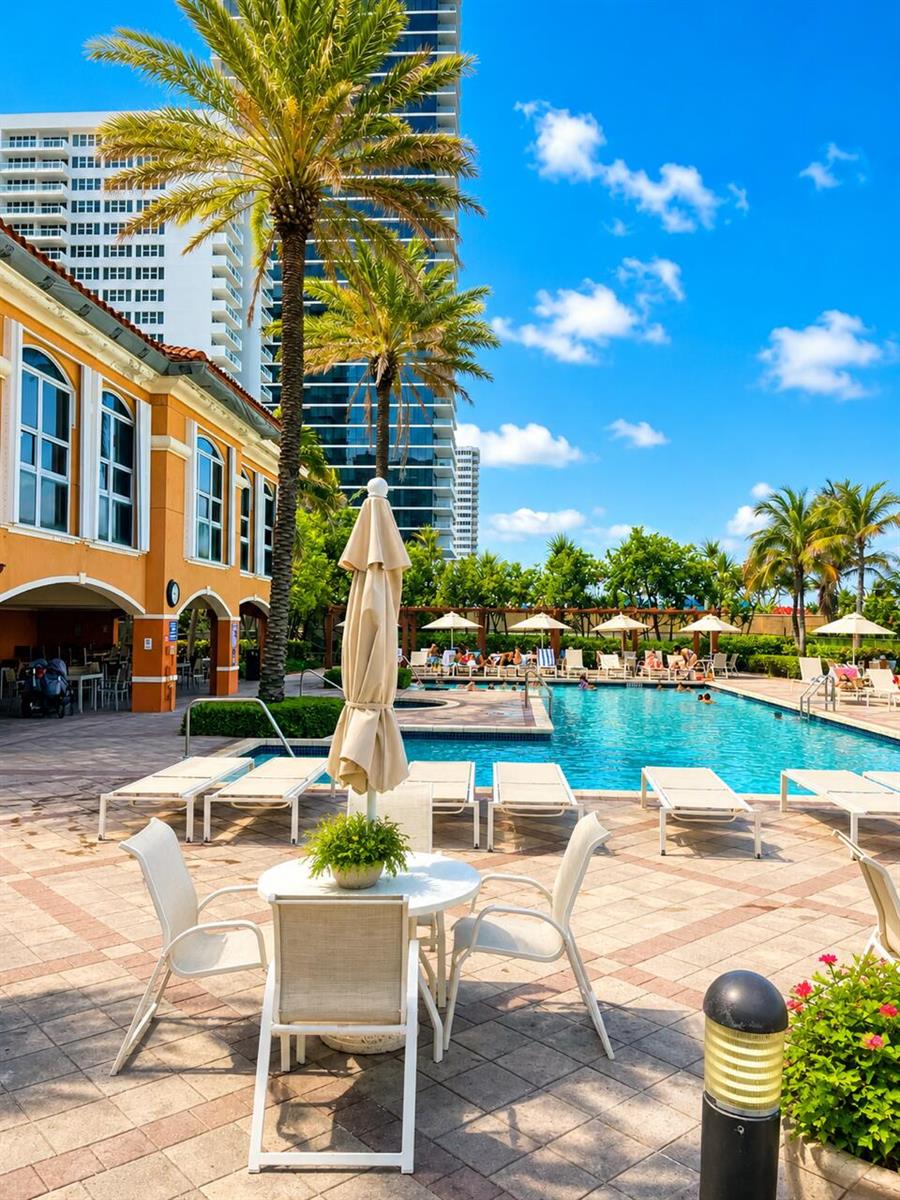 A swimming pool area features lounge chairs, a patio table, and palm trees near buildings — 2080 S Ocean Drive # 505, Hallandale Beach FL 33009, HALLANDALE BEACH, FL