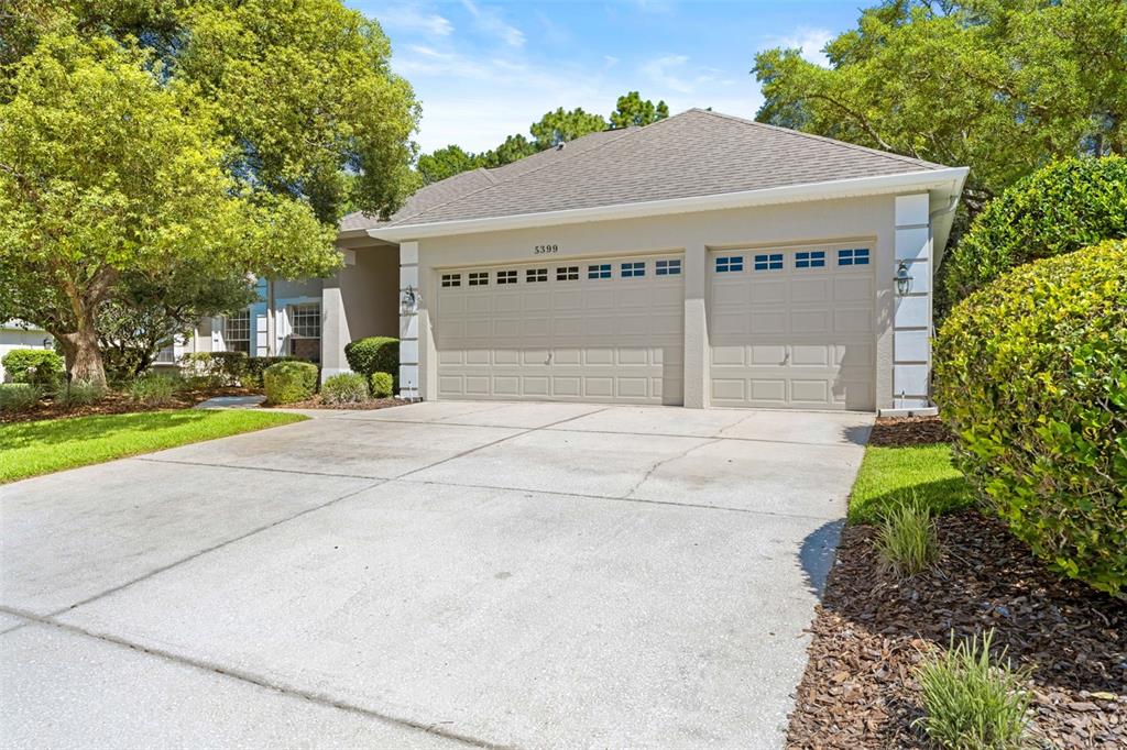 A beige house with a two-car garage and a concrete driveway sits among trees — 5399 CHAMPIONSHIP CUP LANE, SPRING HILL, FL
