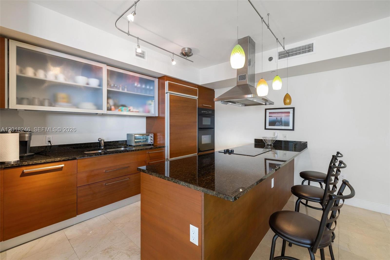 A modern kitchen features wood cabinetry, dark granite countertops, a center island, and bar seating — 1800 S Ocean Dr # 1005, Hallandale Beach FL 33009, HALLANDALE BEACH, FL