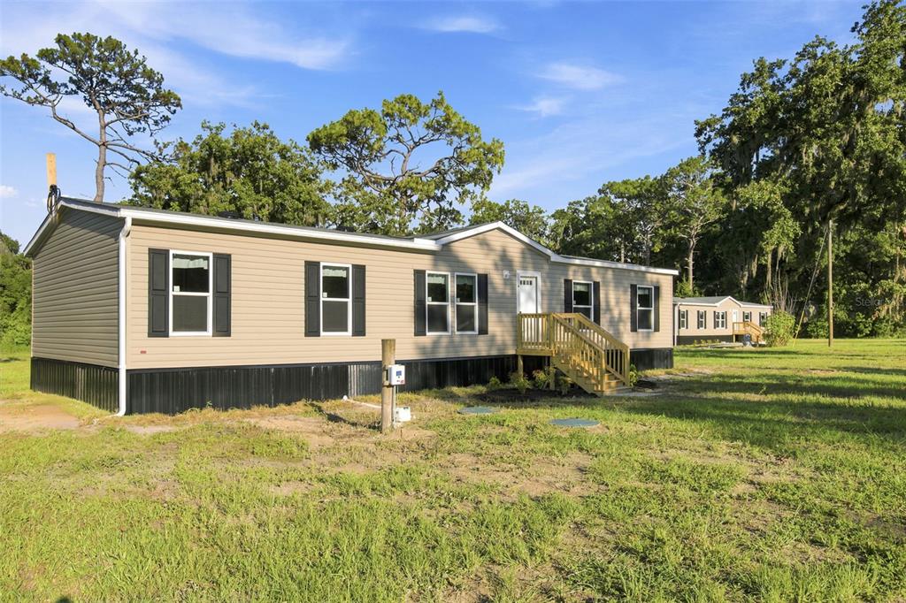 A tan manufactured home with black shutters sits on a grassy lot with trees — 18444 MOORHAVEN DRIVE, SPRING HILL, FL