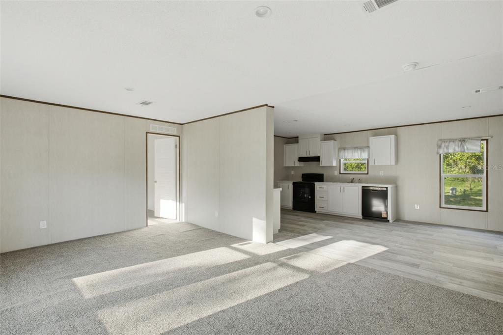 A living room with carpeted flooring transitions into a kitchen featuring white cabinets and appliances — 18444 MOORHAVEN DRIVE, SPRING HILL, FL