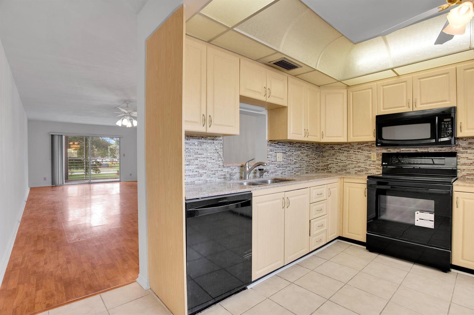 A kitchen with light wood cabinets, black appliances, and tile flooring overlooks an adjacent living area — 2006 Granada Drive # N2, Coconut Creek FL 33066, COCONUT CREEK, FL
