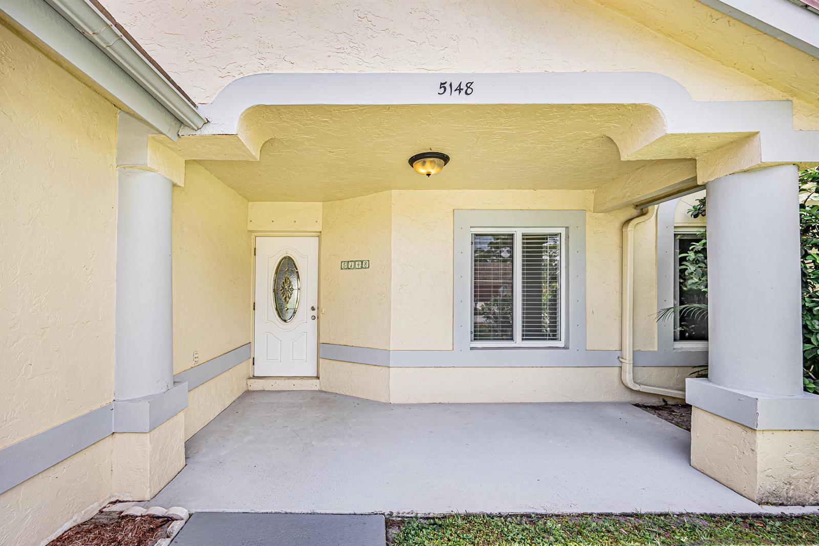 A covered entryway features a white front door, a window, and light yellow stucco walls — 5148 Rosen Boulevard, Boynton Beach FL 33472, BOYNTON BEACH, FL