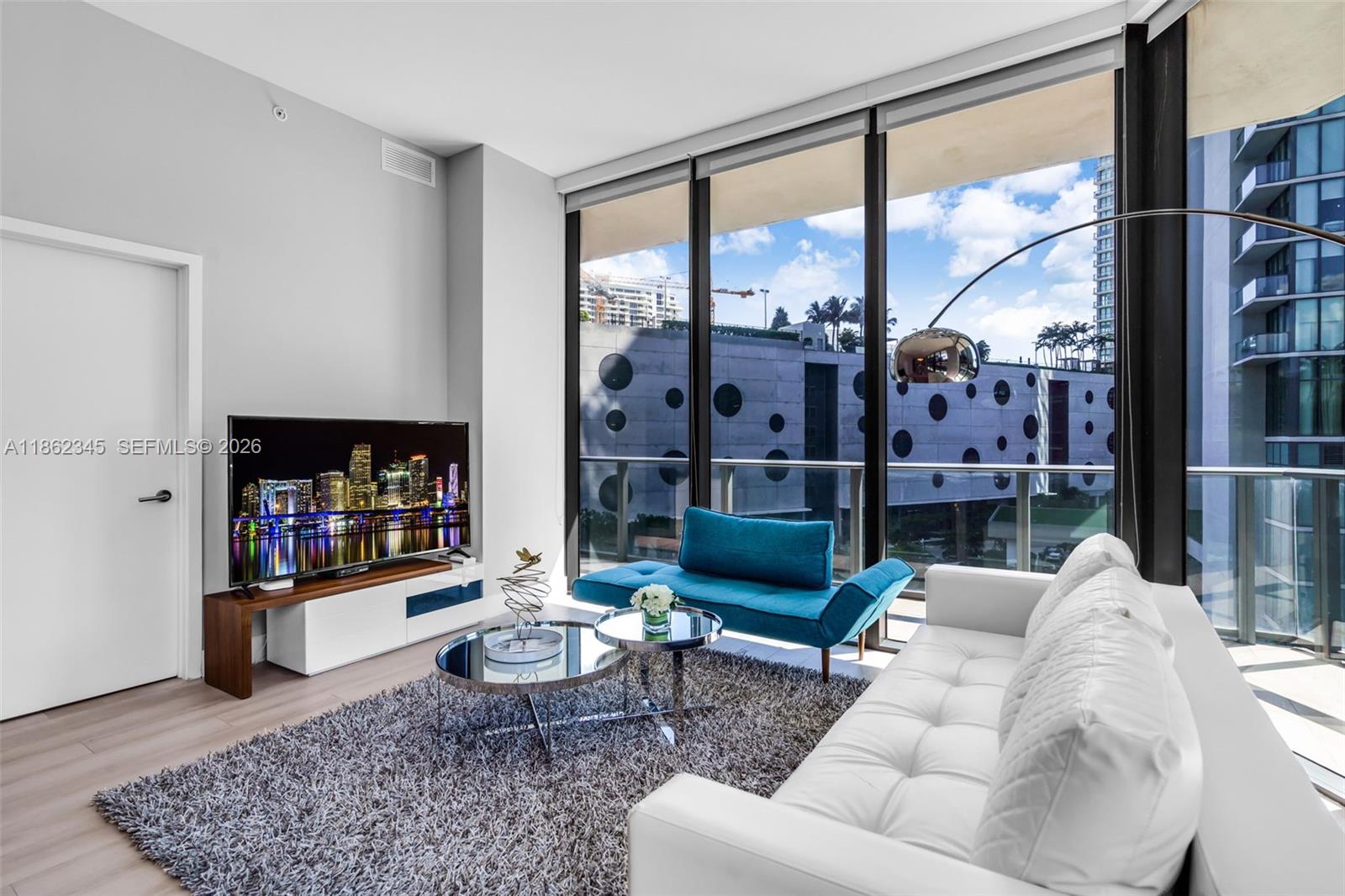 A modern living room features a white sofa, blue chaise, and floor-to-ceiling windows overlooking buildings — 501 NE 31st St # 306, Miami FL 33137, MIAMI, FL