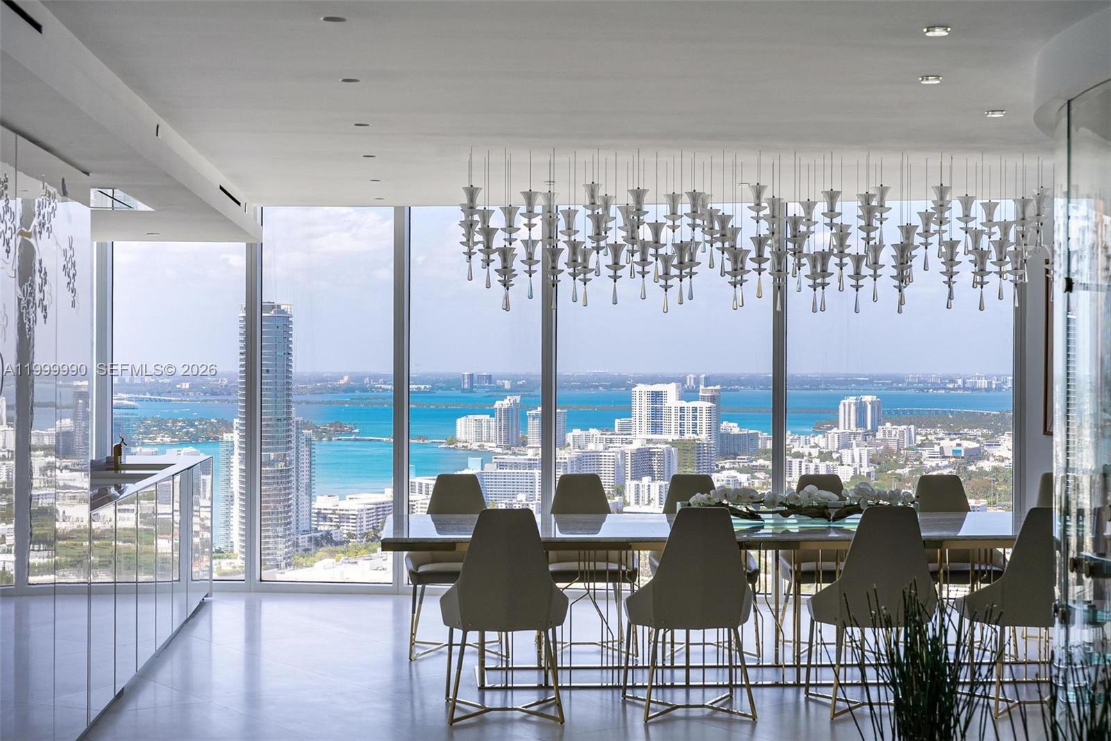 A modern dining room features a large table, sculptural chandelier, and floor-to-ceiling windows overlooking water — 300 S Pointe Dr # 5, Miami Beach FL 33139, MIAMI BEACH, FL