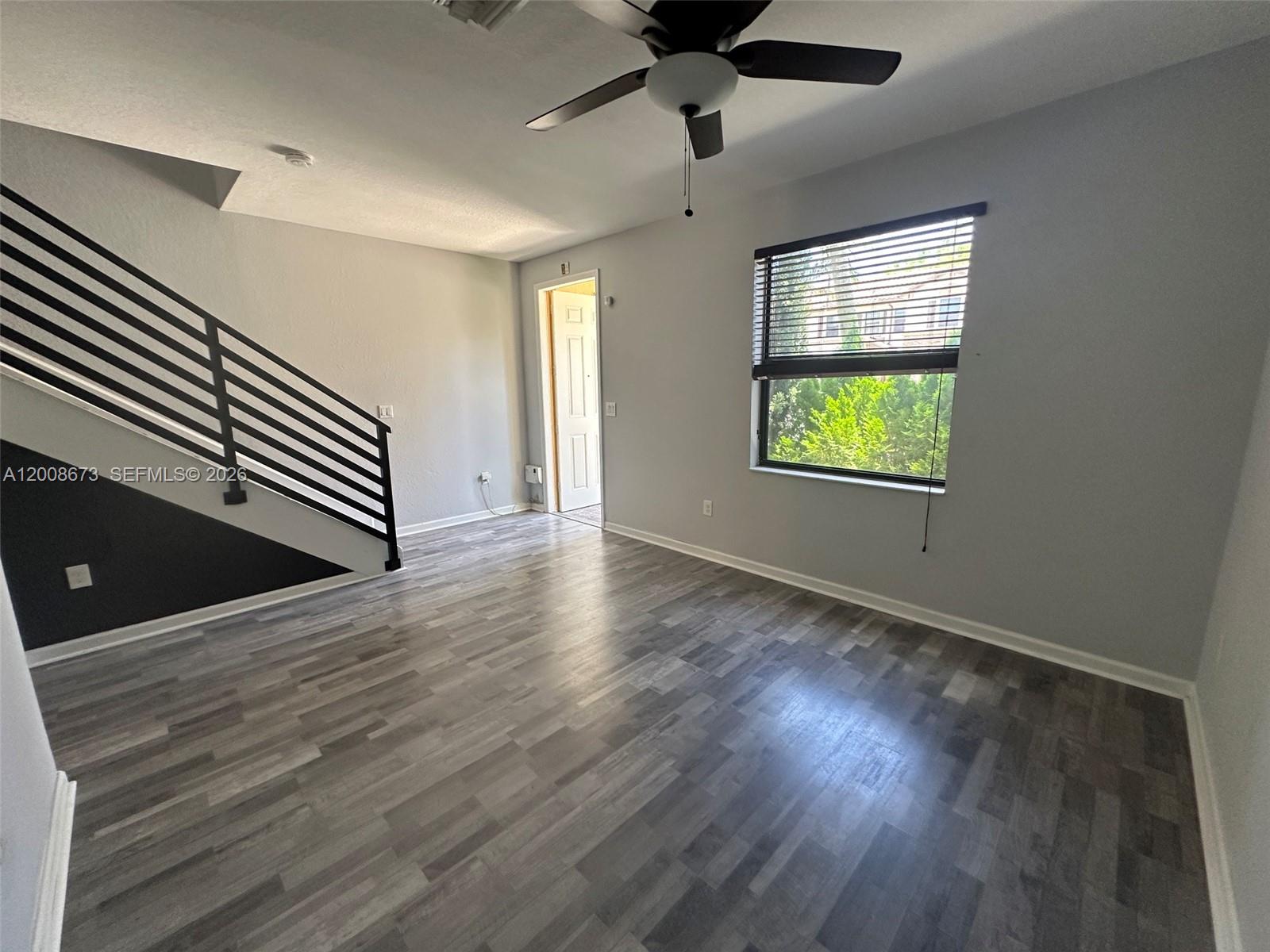 An empty room features gray wood-look flooring, a ceiling fan, and a black-railed staircase — 3211 SE 6th St, Homestead FL 33033, HOMESTEAD, FL