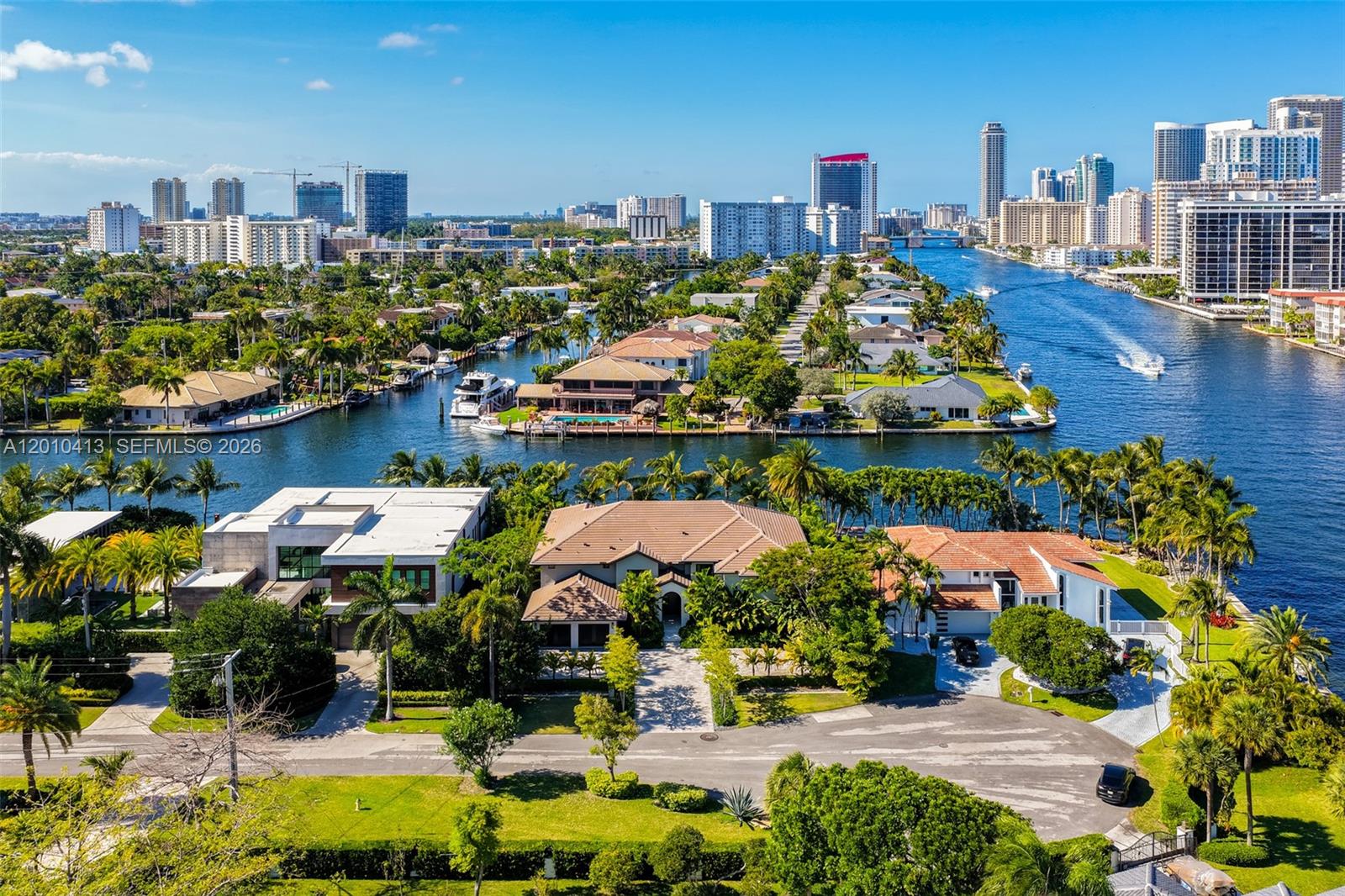 An aerial view shows waterfront homes along a canal with a city skyline in the background — 110 Holiday Dr, Hallandale Beach FL 33009, HALLANDALE BEACH, FL