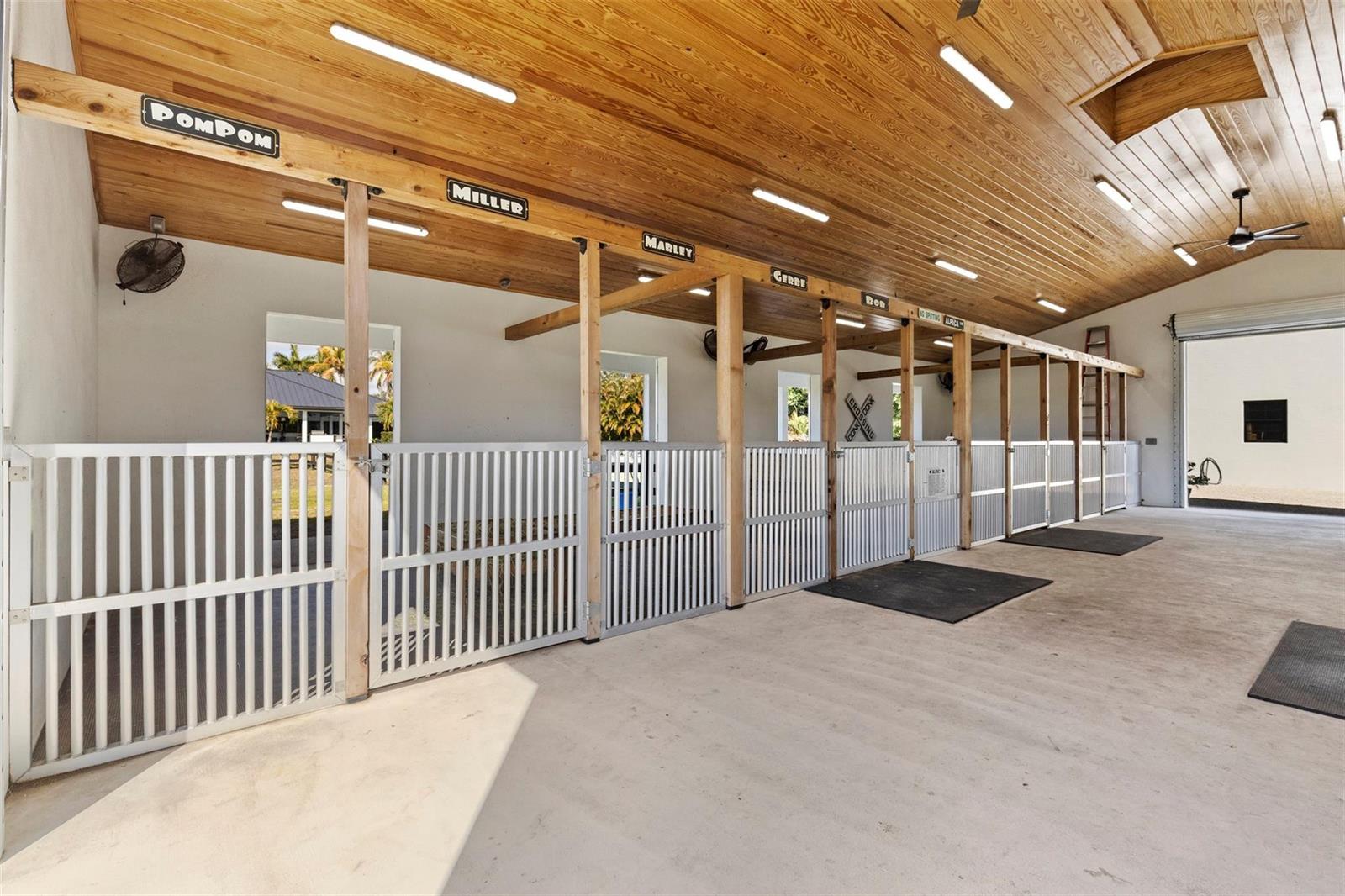 A horse stable interior features multiple stalls with white metal gates and wood-paneled ceilings — 14473 Draft Horse Lane, Wellington FL 33414, WELLINGTON, FL