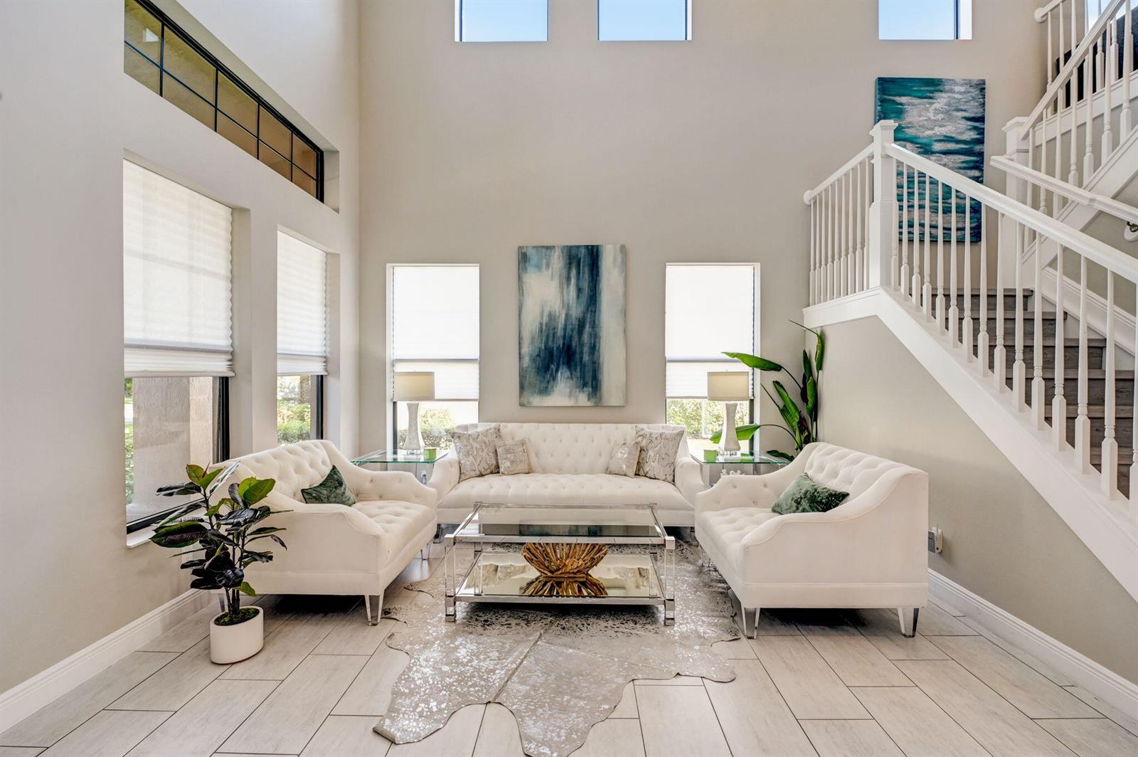 A bright living room features white tufted furniture, a glass coffee table, and a staircase — 16050 Tuscany Estates Drive, Delray Beach FL 33446, DELRAY BEACH, FL