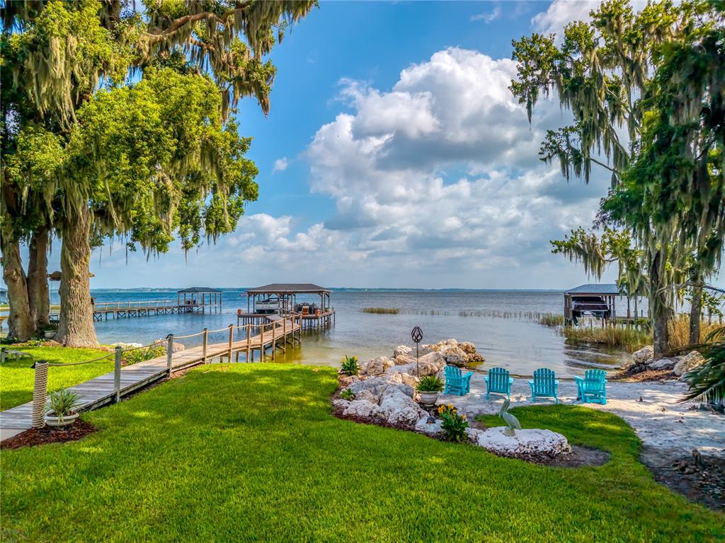 A grassy backyard features a wooden dock, boat slips, and four blue chairs by the water — 7311 BIRDS NEST COURT, YALAHA, FL