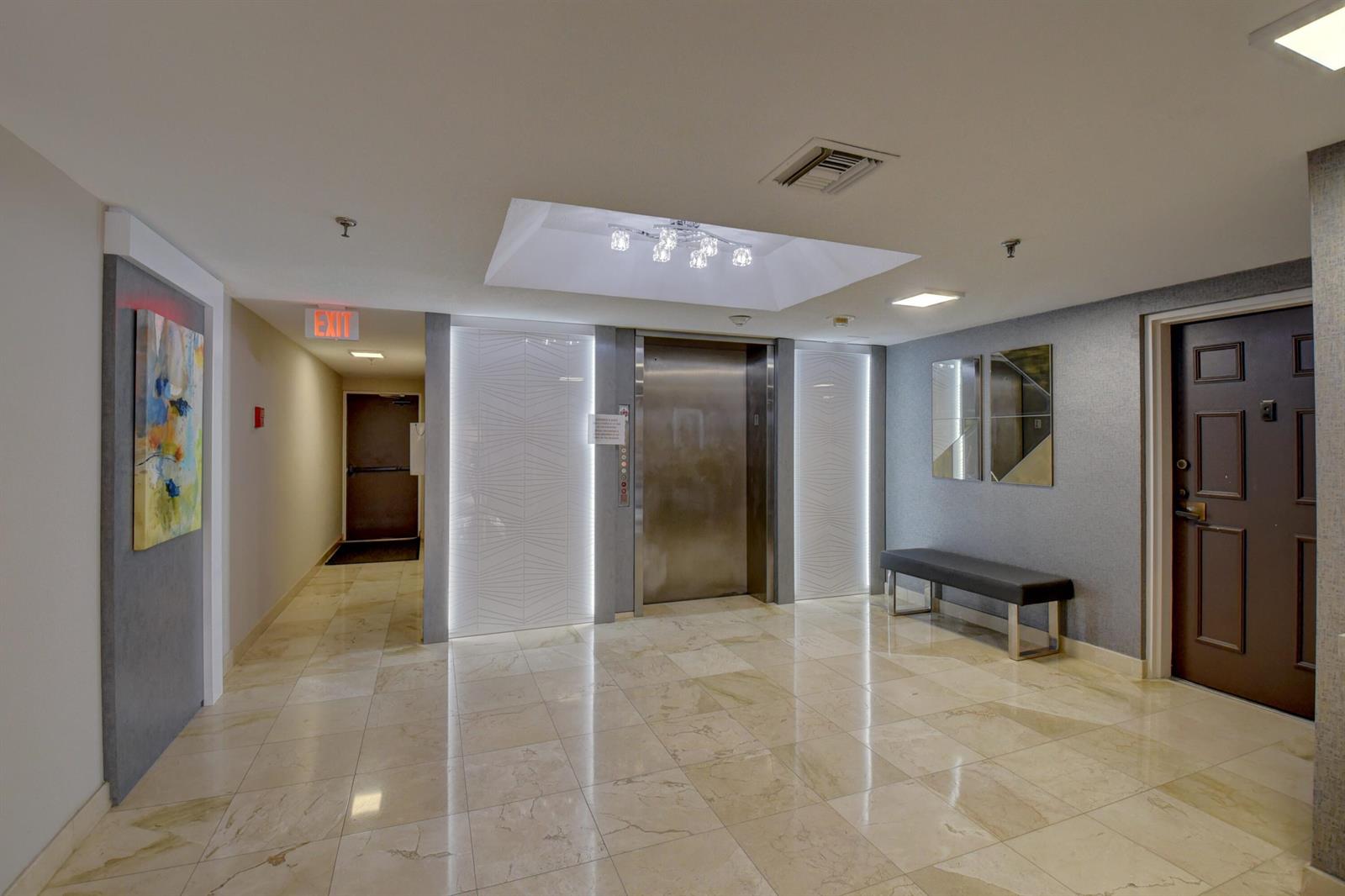 A modern elevator lobby features marble floors, a metallic elevator door, and a dark bench — 7194 Promenade Drive # 202, Boca Raton FL 33433, BOCA RATON, FL