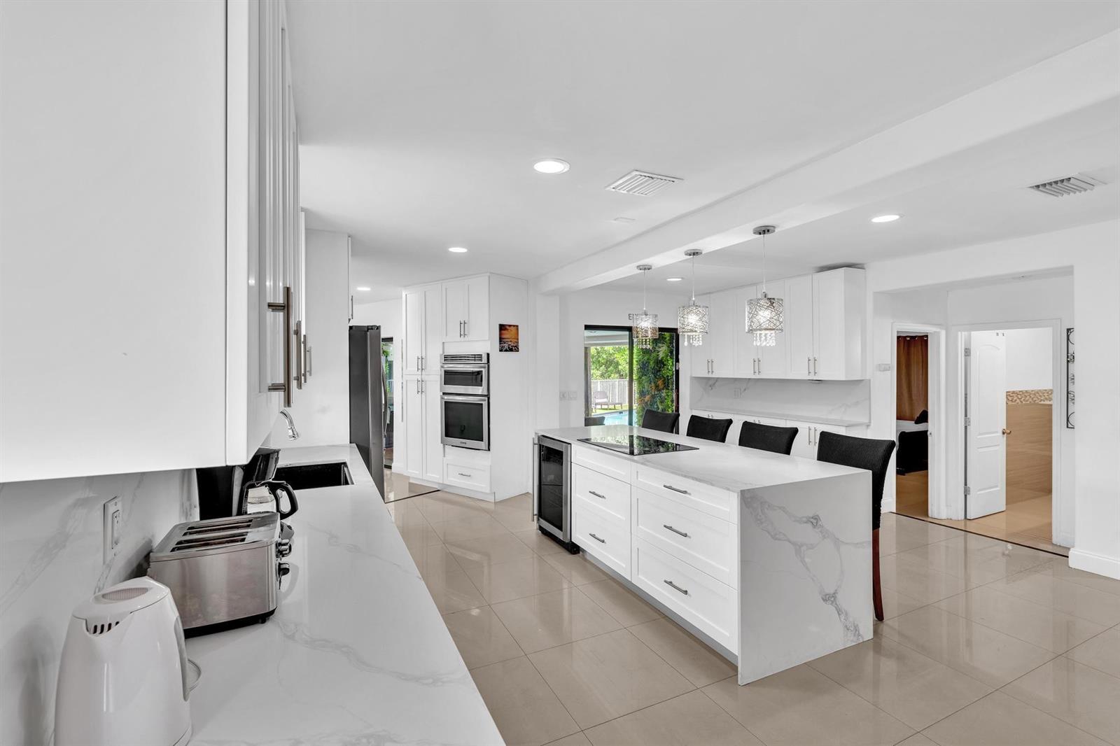 A modern white kitchen features a marble-topped island, stainless steel appliances, and tile flooring — 5901 W Broward Boulevard, Plantation FL 33317, PLANTATION, FL