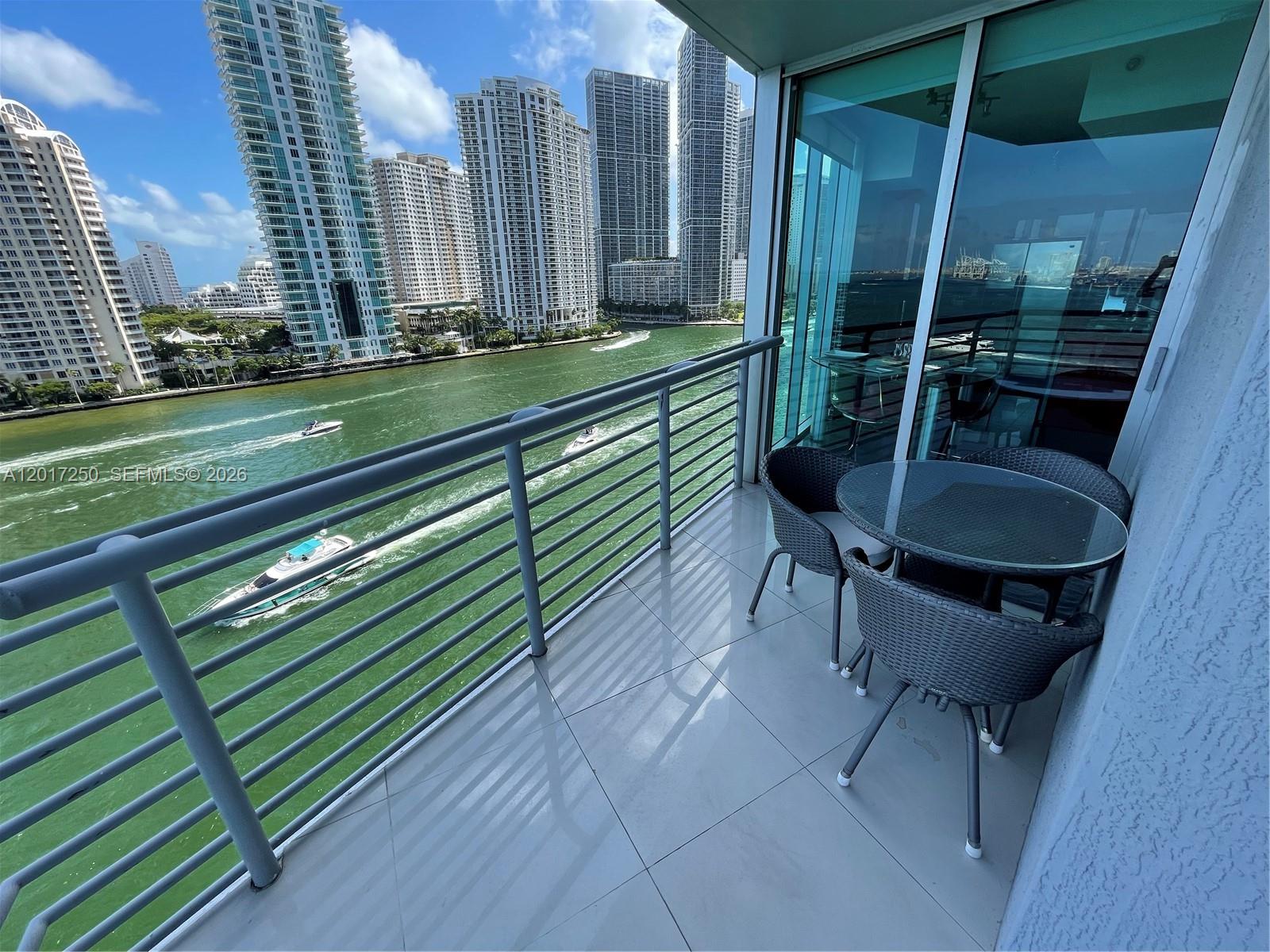 A balcony with a glass-topped table and chairs overlooks a river and city skyline — 335 S Biscayne Blvd # 1209, Miami FL 33131, MIAMI, FL
