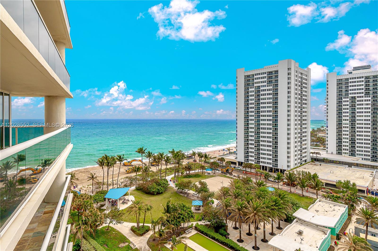An elevated view from a balcony overlooking a beach, palm trees, and high-rise buildings — 1850 S Ocean Dr # 1105, Hallandale Beach FL 33009, HALLANDALE BEACH, FL