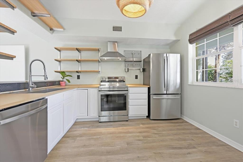 A kitchen features white cabinets, stainless steel appliances, open wood shelving, and light wood flooring — 70 SAN JUAN DRIVE UNIT D101, PALM COAST, FL