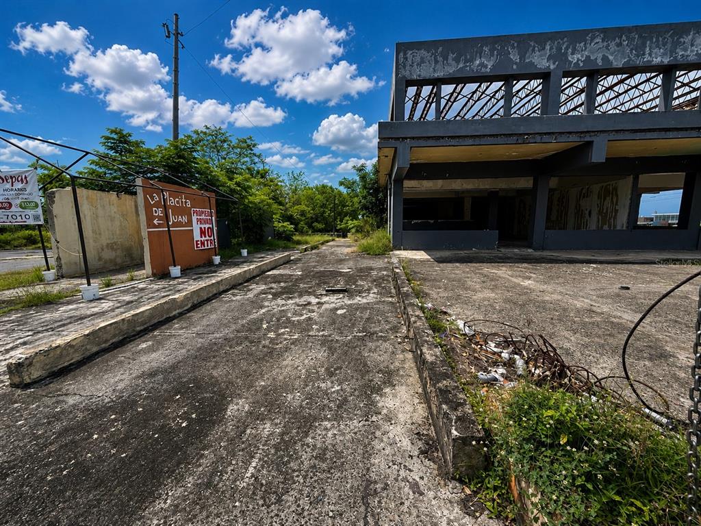 An exterior view of a weathered, abandoned concrete building with a paved driveway and signage — Calle 28 URB. MIRAFLORES, BAYAMON, PR