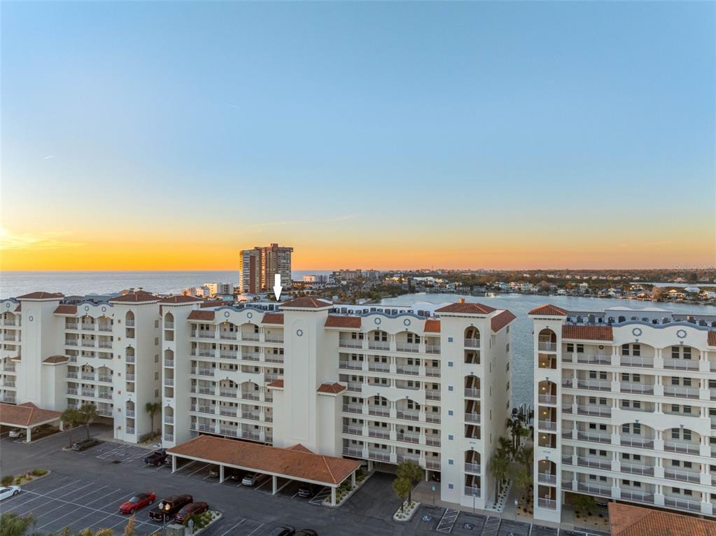An aerial view shows a multi-story waterfront condominium complex with a parking lot at sunset — 17717 GULF BOULEVARD UNIT 702, REDINGTON SHORES, FL