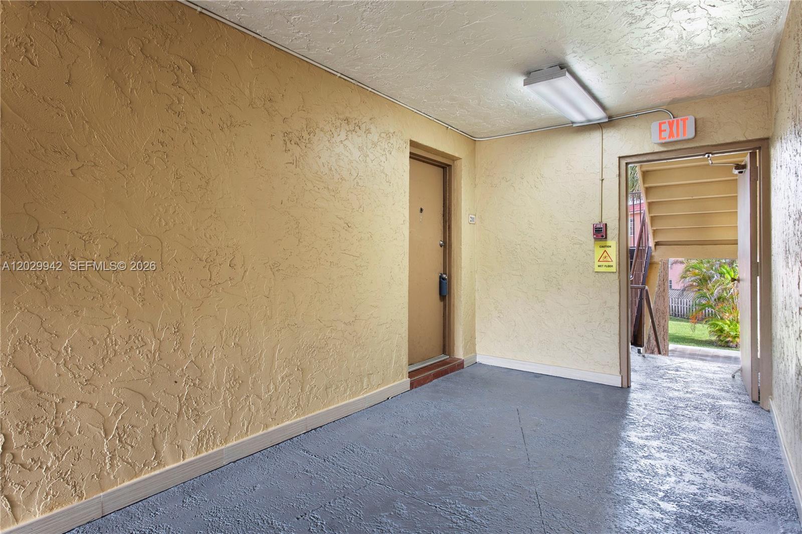 An apartment hallway features textured yellow walls, a grey floor, and an open exit door — DORAL, FL