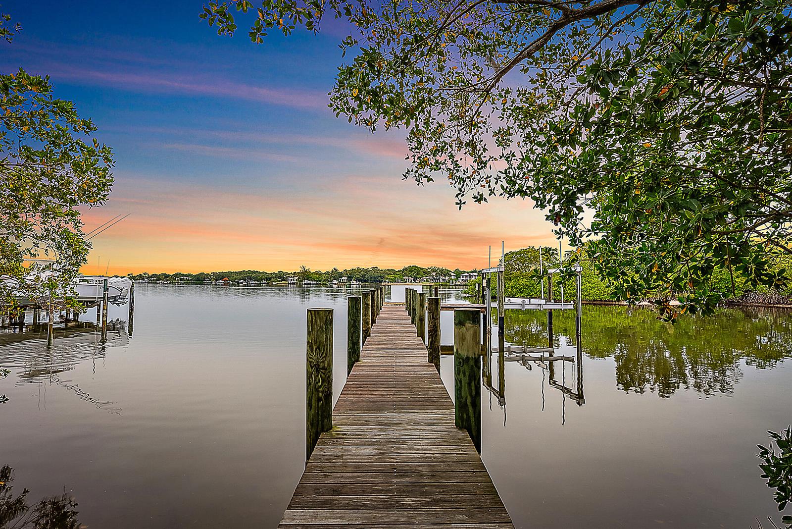 A wooden dock extends into a calm waterway at sunset, framed by overhanging tree branches — 18189 49th Terrace, Jupiter FL 33458, JUPITER, FL